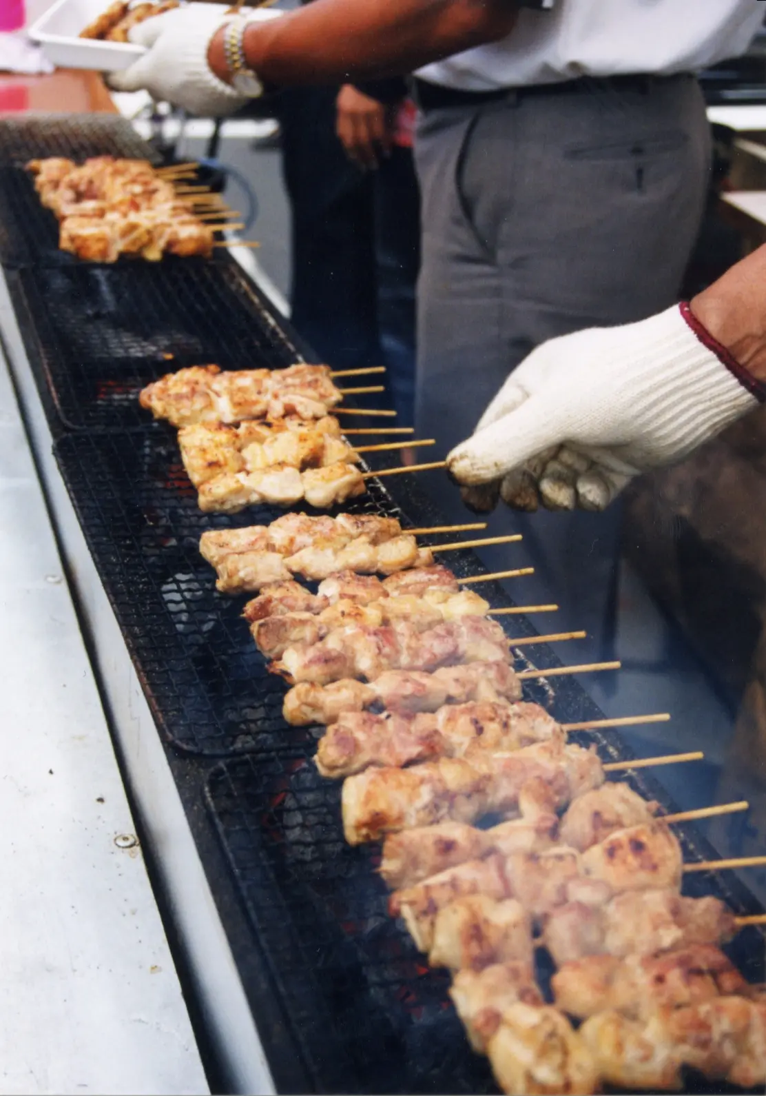 A cook turning rows of chicken skewers over a long charcoal grill